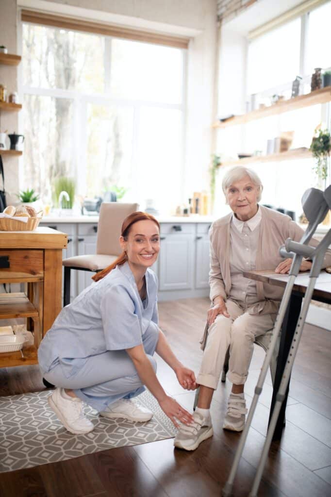 Caregiver smiling while helping aged woman to lace shoes